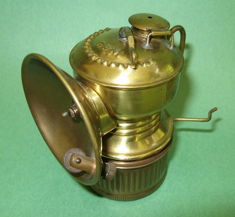 Gem Brass LSide.jpg - GEM BRASS - Brass lamp marked GEM in raised letters on top, with hook and cap braces, 3.5 in. to water door, 2 ½ in dia. brass reflector, mfg. by GrierBros., unfired condition; ex-Bob Schroth collection  (The Grier Bros. Co. was founded in 1839 in Pittsburgh, PA to manufacture miner's equipment.  By the 1880s the company was very active in the manufacture and sales of oil wick lamps with branches in Hancock, MI and Dubois, PA.  They produced  a line of face and driver's wick lamps for both oil and Sunshine fuels including the popular STAR brand in a shield trademark (check out the Grier lamps in the oilwick lamp photos).  As wick lamps started phasing out with the increased use of carbide lamps, the company started producing carbide lamp models perhaps as early as 1910.  They marketed at least 26 different numbered carbide cap lamp models.  In 1924, the Grier Bros. plant and product line was purchased by the Gem Manufacturing Co. and a year later a product line of carbide lamps with the GEM product name as shown here was introduced that led to at least 18 different GEM lamp models being advertised; see Pohs, Miner's Flame Light, pp 245, 429-434)